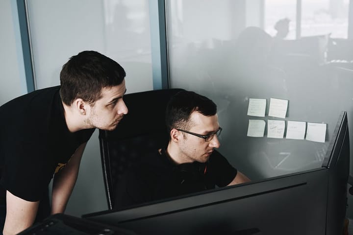 Two people in black shirts are working at a desk, one standing and peering over the shoulder of the other, who is seated on a